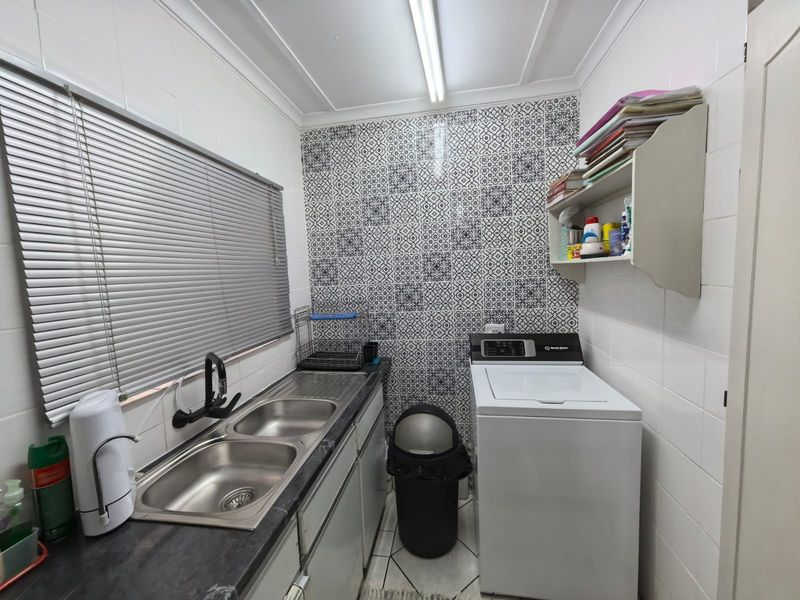 A functional laundry room featuring a double stainless steel sink, dark laminate countertop, patterned feature wall tiles, and built-in storage shelving.
