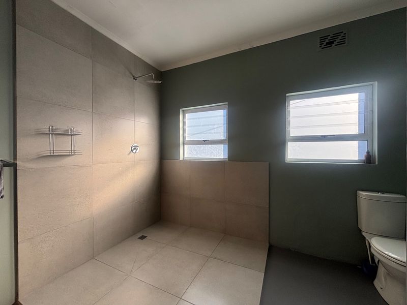 A clean, modern bathroom featuring a spacious walk-in shower with large format tiling, a toilet, and dual louver windows for natural light and ventilation.