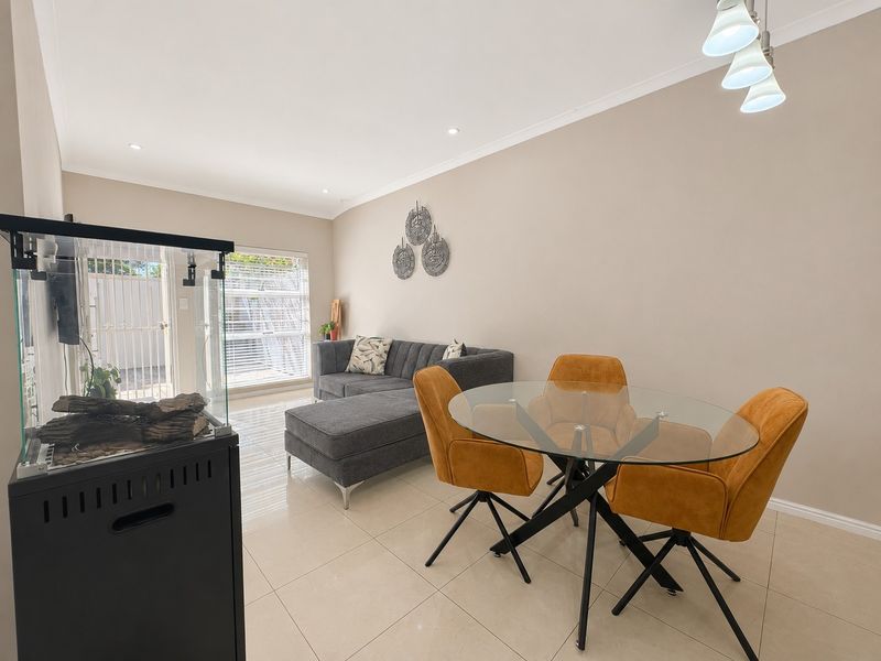 An open-plan living and dining area featuring neutral tiled flooring, recessed lighting, and access to an outdoor space through a secure gate.