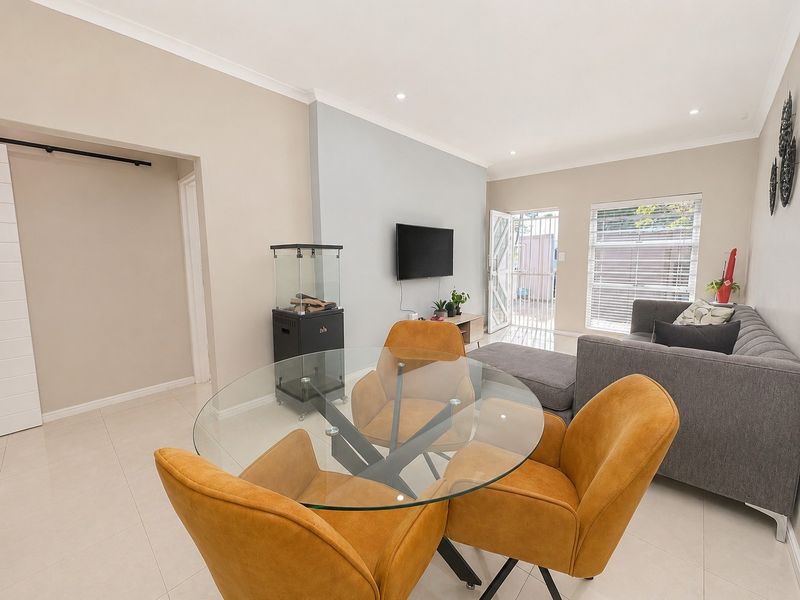 A bright open-plan living and dining area featuring neutral wall tones, tiled flooring, recessed lighting, and direct access to the exterior via a security gate.