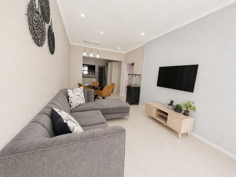 An open-plan living and dining area featuring neutral tiled flooring, recessed downlighting, and a wall-mounted television, leading through to a kitchen space.