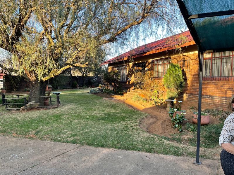 A view of the rear brick facade of the house, featuring a well-maintained garden, mature trees, and a concrete driveway area.