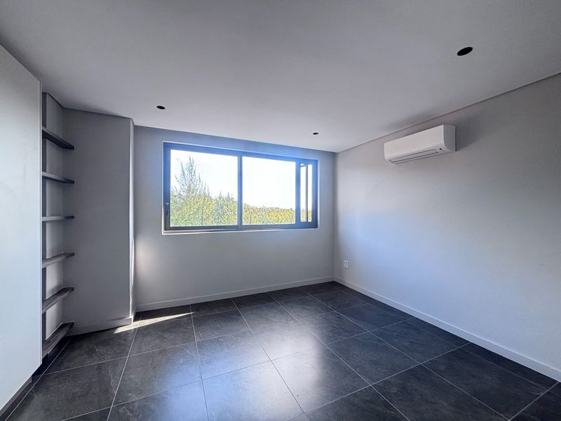 A bright, modern, and empty bedroom featuring dark tiled flooring, built-in shelving, and a wall-mounted air conditioning unit.