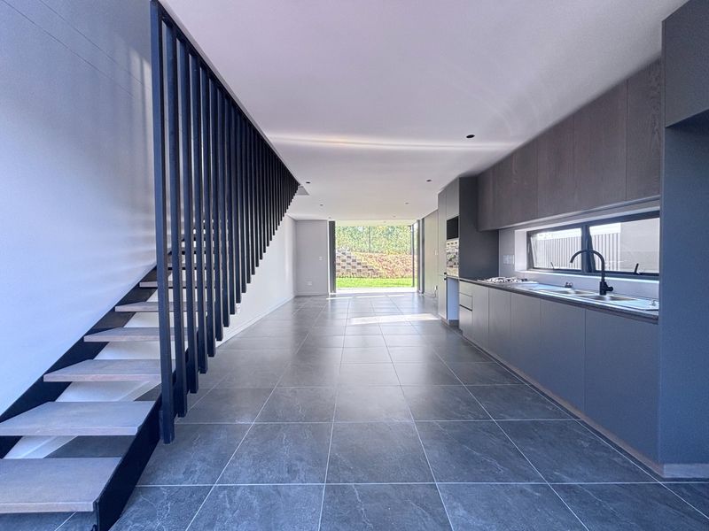 Modern open-plan living area featuring a sleek kitchen with built-in appliances, tiled flooring, and a contemporary staircase with a striking vertical balustrade.