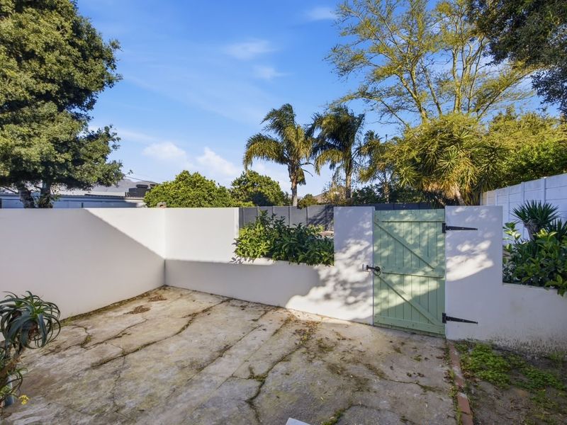A private rear courtyard area featuring concrete paving, a boundary wall, and a timber access gate, surrounded by established garden greenery.