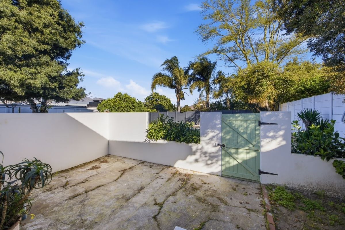 A private rear courtyard area featuring concrete paving, a boundary wall, and a timber access gate, surrounded by established garden greenery.