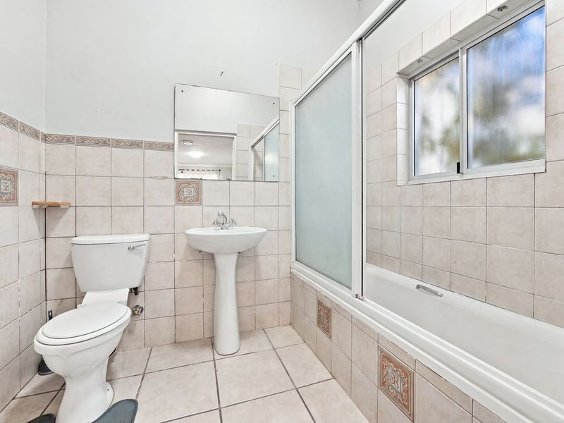 A functional bathroom featuring a pedestal basin, toilet, and a bathtub with a sliding shower screen, finished with neutral ceramic wall and floor tiles.