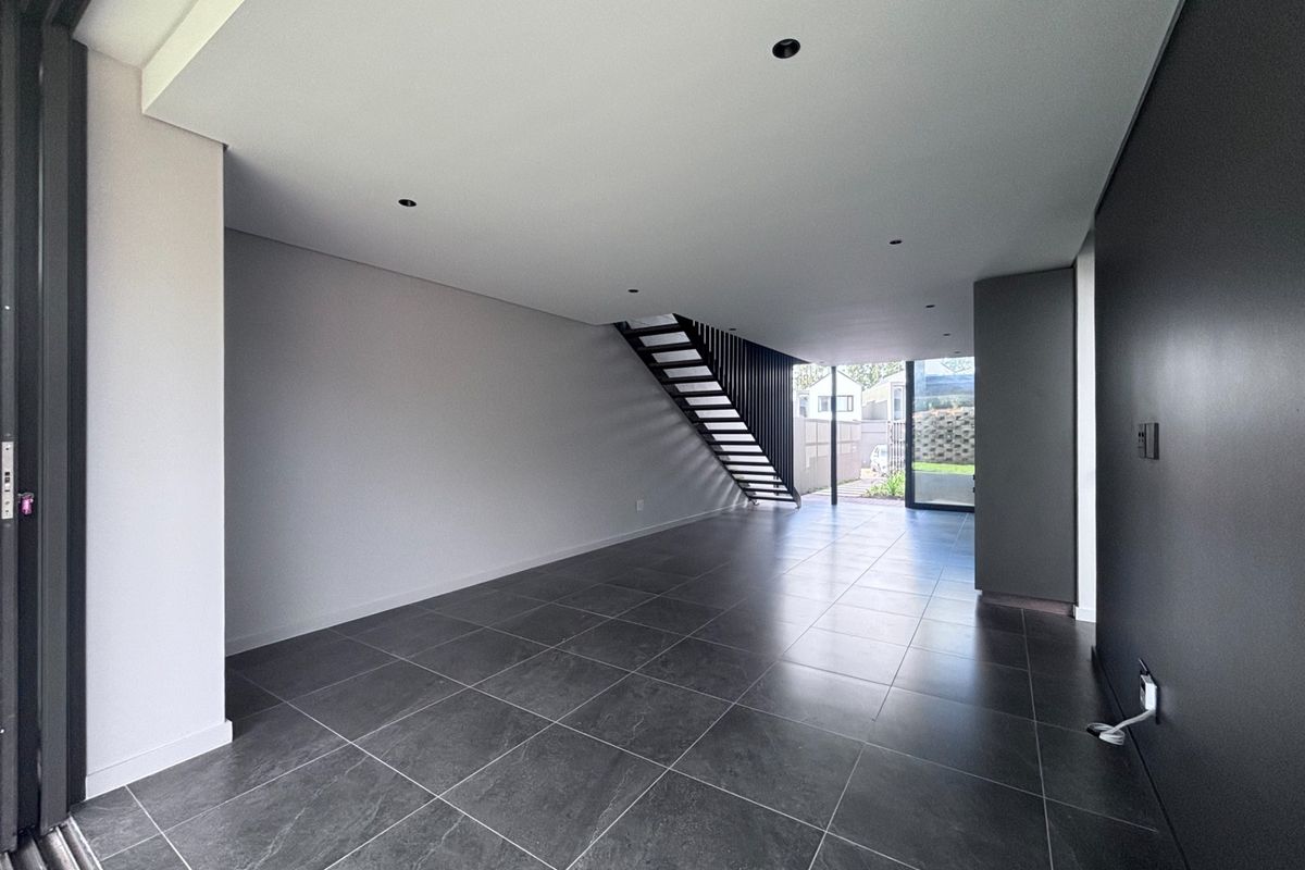 A modern, open-plan living area featuring large format floor tiles, recessed lighting, and a sleek floating staircase leading to the upper level.