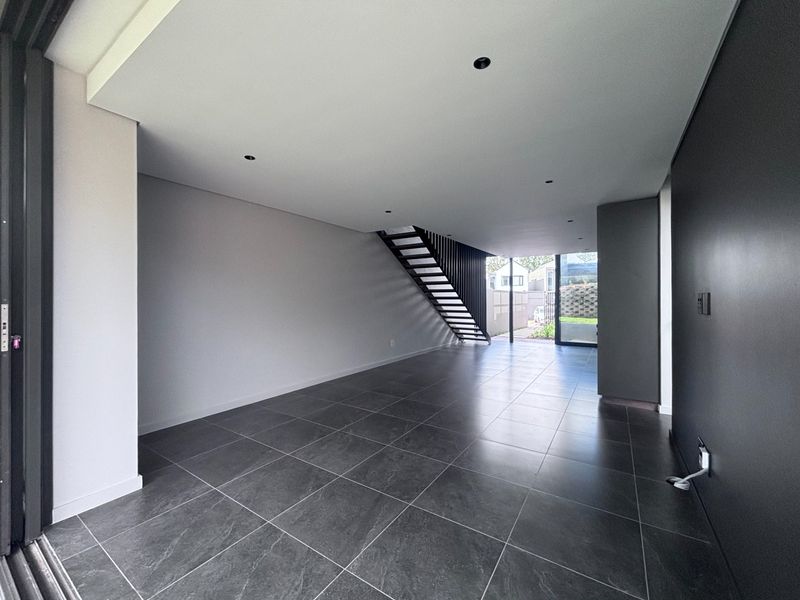 A modern, open-plan living area featuring large format floor tiles, recessed lighting, and a sleek floating staircase leading to the upper level.