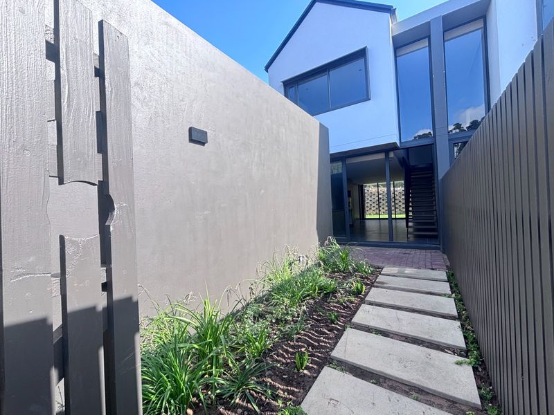 A modern townhouse entrance featuring a paved walkway, landscaped garden bed, and contemporary architectural facade with large glass windows.