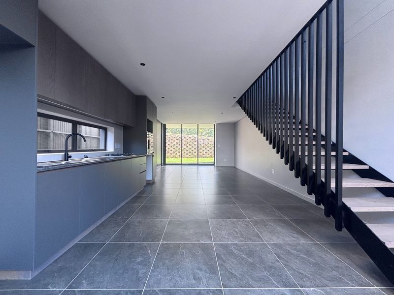 Modern open-plan living area featuring a sleek kitchen with dark cabinetry, stone countertops, and a striking staircase, leading to a bright rear garden view.