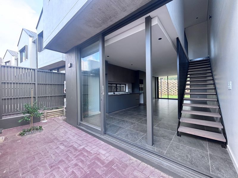 Modern open-plan living area with sleek kitchen cabinetry, tiled floors, and a staircase, seamlessly connecting to a private paved patio through large sliding doors.