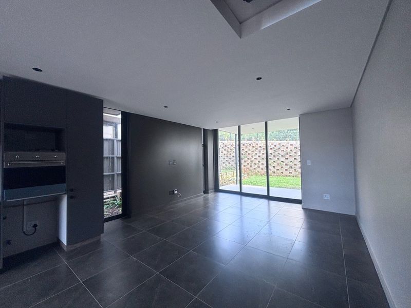 Modern open-plan living area featuring dark cabinetry, built-in oven, large format floor tiles, and sliding glass doors leading to a private garden.