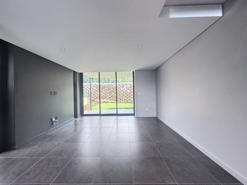 A modern, empty living room featuring large format floor tiles, recessed lighting, and sliding glass doors providing access to the rear garden.