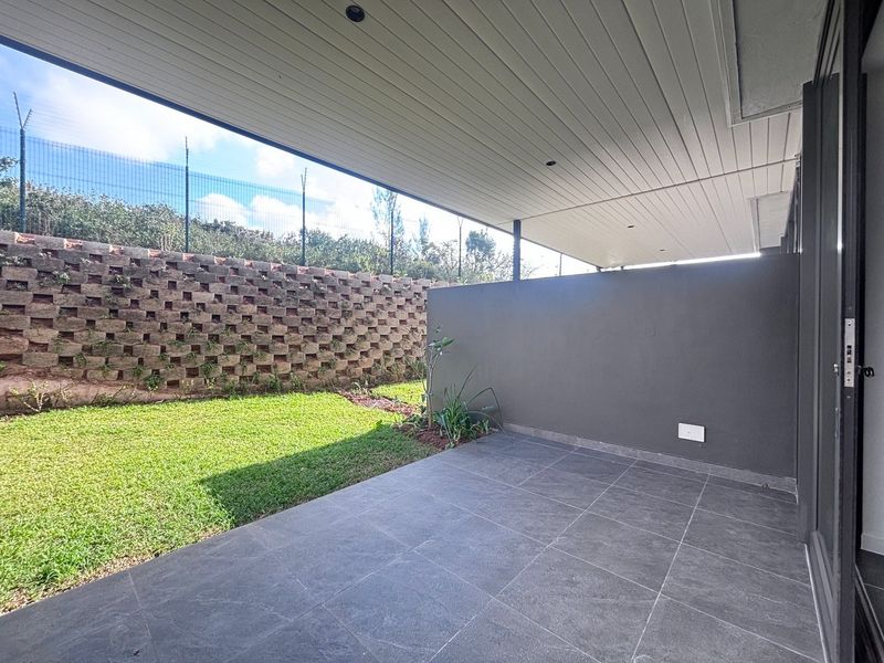A covered patio area featuring tiled flooring and recessed lighting, overlooking a private garden with a retaining wall and perimeter fencing.