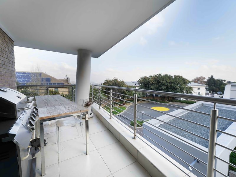 A spacious covered balcony featuring tiled flooring and a stainless steel balustrade, offering an elevated view of the surrounding complex.