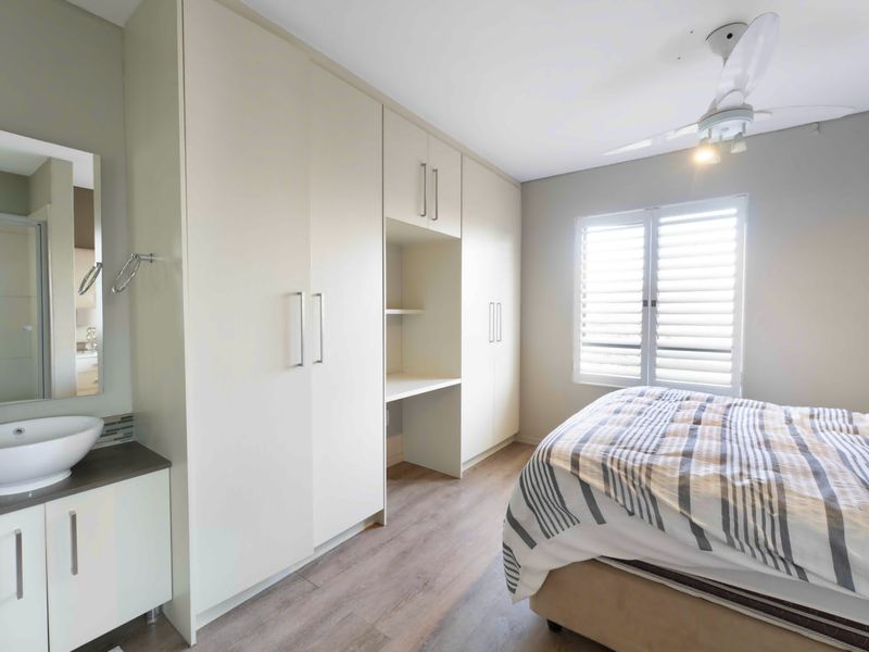 A well-appointed bedroom featuring built-in cabinetry with an integrated desk, laminate flooring, plantation shutters, and a convenient vanity basin area.