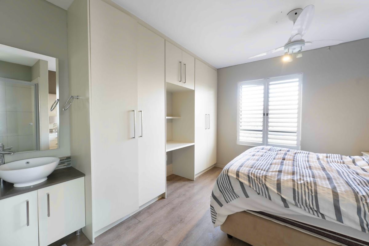 A well-appointed bedroom featuring built-in cabinetry with an integrated desk, laminate flooring, plantation shutters, and a convenient vanity basin area.