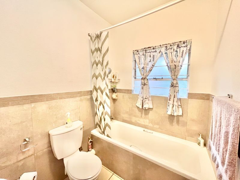 A functional bathroom featuring neutral wall and floor tiling, a standard bathtub with shower curtain, and a louvered window for ventilation.