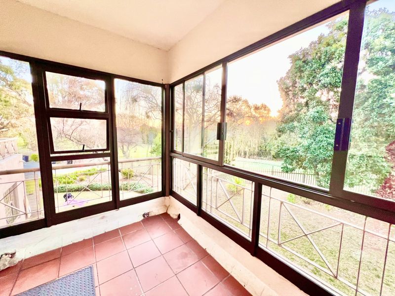 An enclosed balcony featuring tiled flooring and large sliding glass windows that offer expansive views of the surrounding garden and trees.