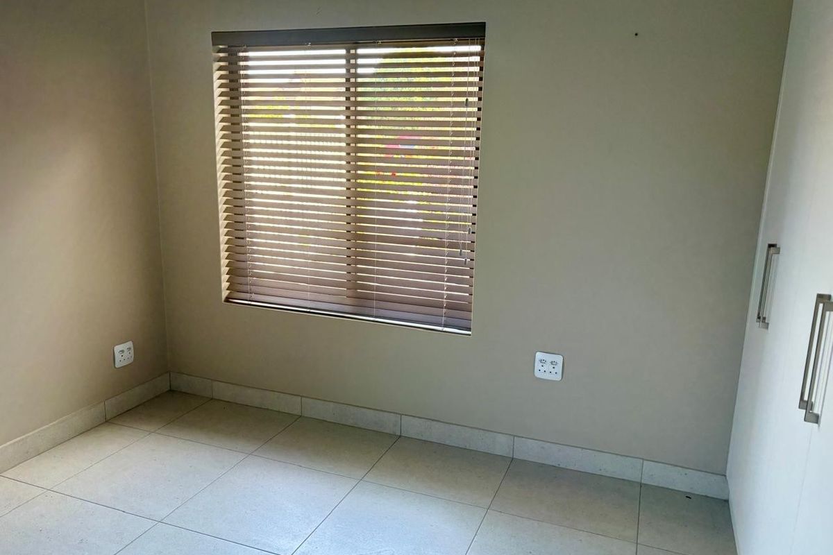 A clean, empty bedroom featuring neutral tiled flooring, built-in cupboards, and venetian blinds on the window.