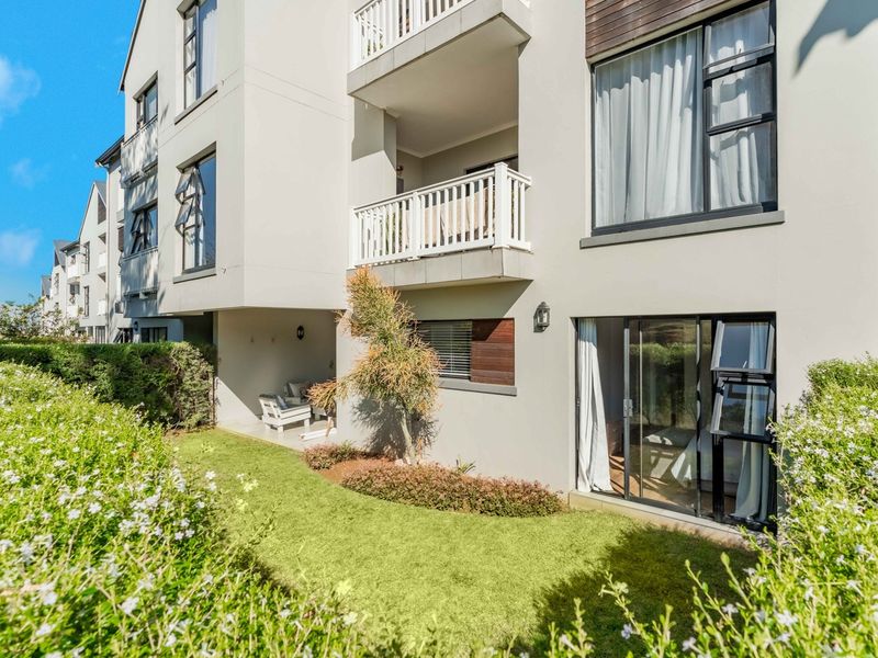 Rear view of a modern apartment building featuring private balconies, sliding glass doors, and a well-maintained ground-floor garden area.