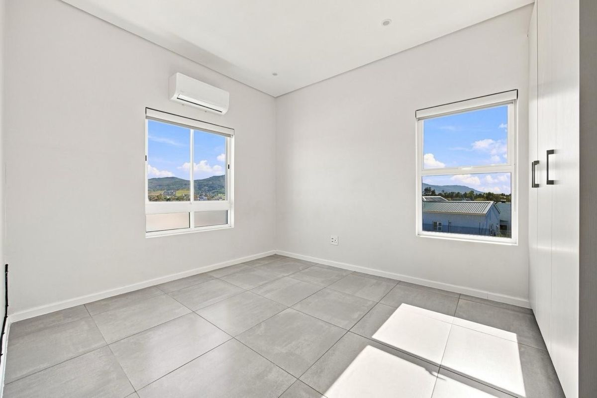 A bright, modern, and empty bedroom featuring large windows, tiled flooring, built-in cupboards, and a wall-mounted air conditioning unit.