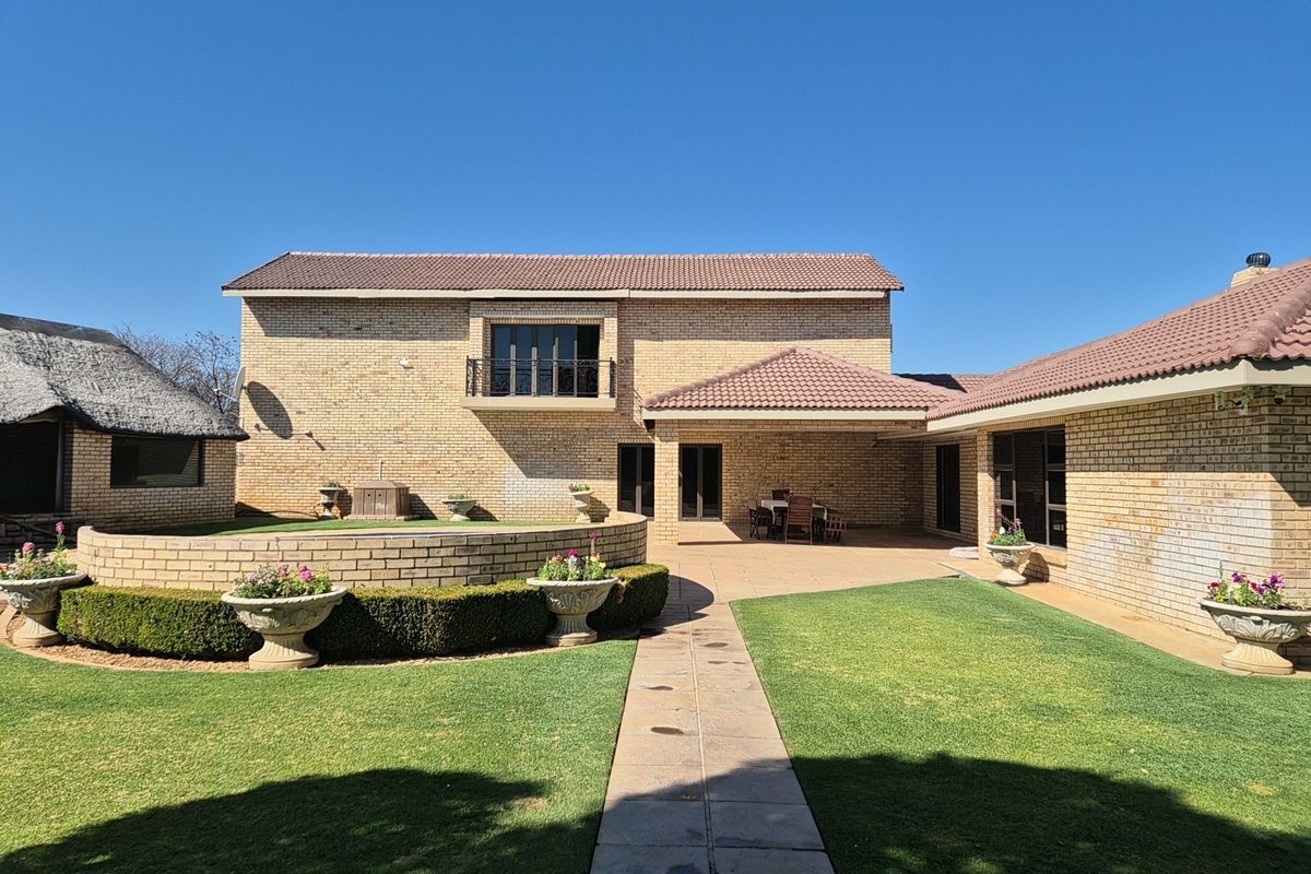 Rear view of a double-story brick house featuring a covered patio, balcony, landscaped garden with a retaining wall, and a paved walkway.