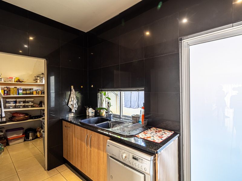 A functional scullery featuring dark wall tiles, wood-finish cabinetry, a double sink, and an adjoining walk-in pantry with ample storage shelving.