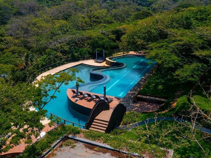 An aerial view of a large, uniquely shaped swimming pool surrounded by lush forest, featuring a pedestrian bridge and wooden decking.