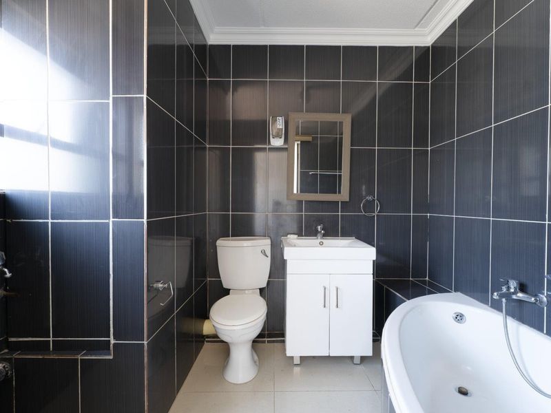 A compact bathroom featuring dark wall tiles, a white vanity with basin, a toilet, and a bathtub with a shower mixer.