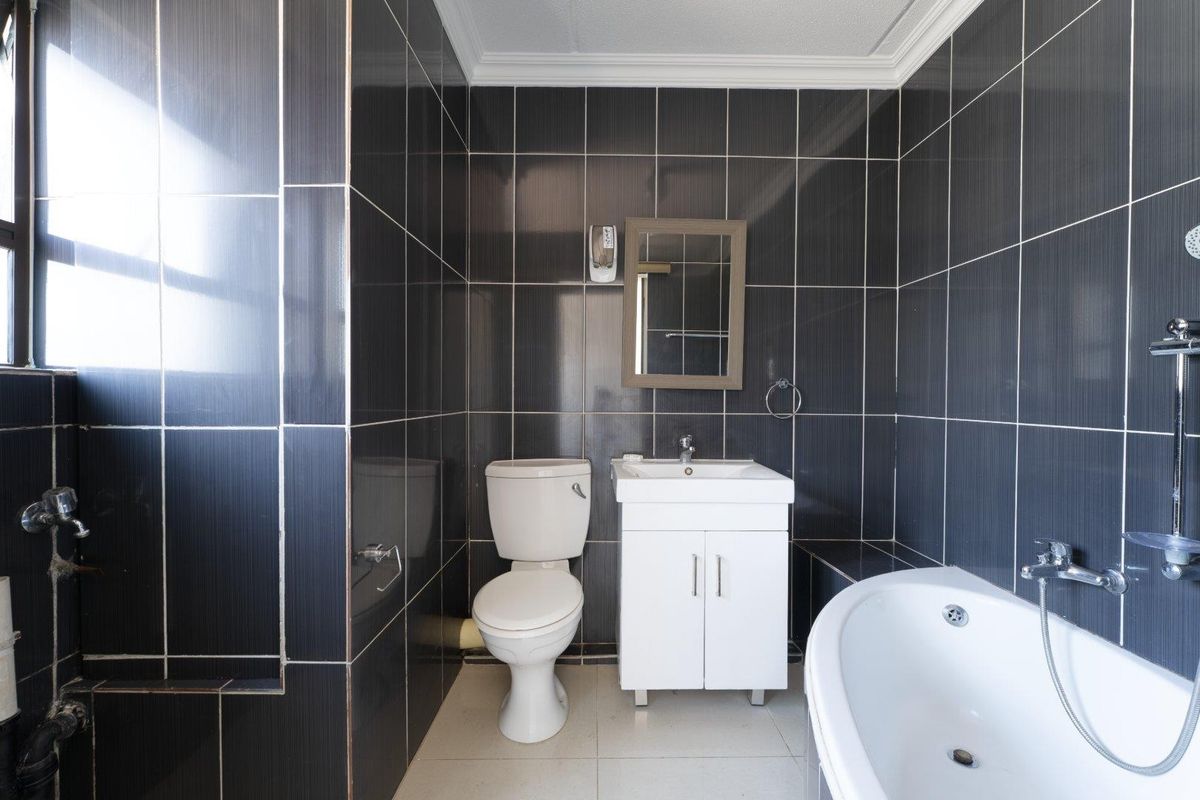 A compact bathroom featuring dark wall tiles, a white vanity with basin, a toilet, and a bathtub with a shower mixer.