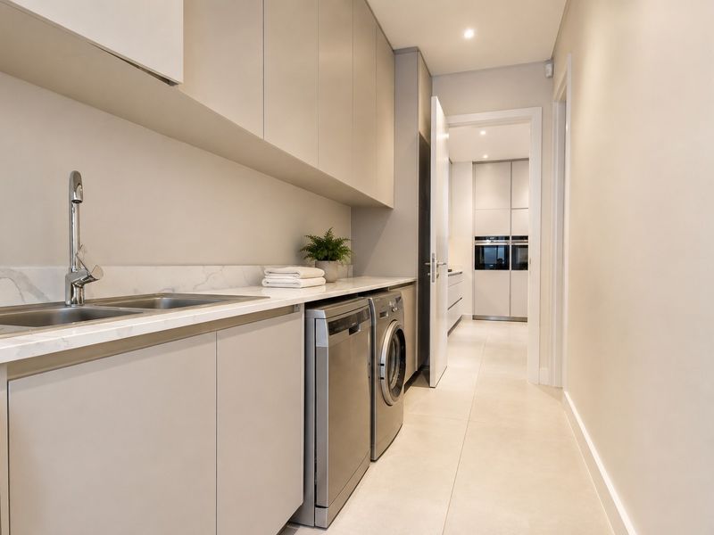 Modern, well-appointed laundry room featuring sleek built-in cabinetry, stone countertops, a stainless steel sink, and integrated appliance spaces with neutral tiled flooring.
