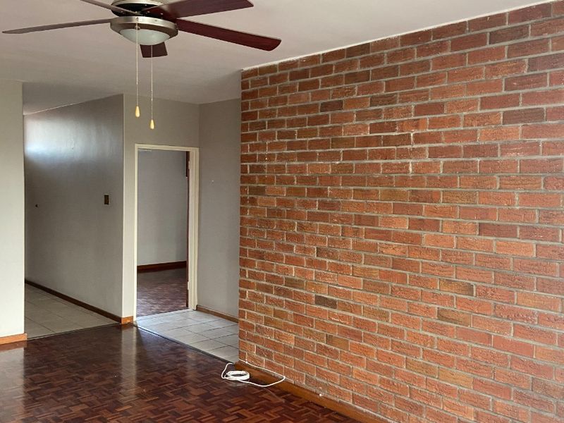 Spacious living area featuring classic parquet flooring, a prominent exposed brick feature wall, and a ceiling fan, leading into a tiled hallway.