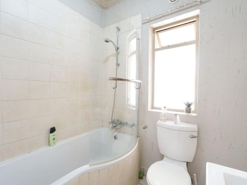 A compact bathroom featuring a curved bathtub with a glass shower screen, a standard toilet, and a top-hung window with tiled wall finishes.