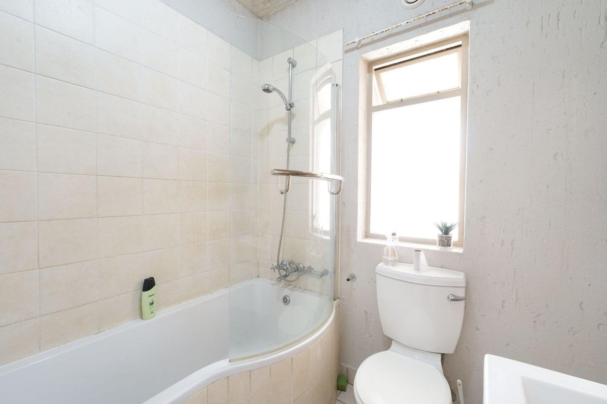 A compact bathroom featuring a curved bathtub with a glass shower screen, a standard toilet, and a top-hung window with tiled wall finishes.