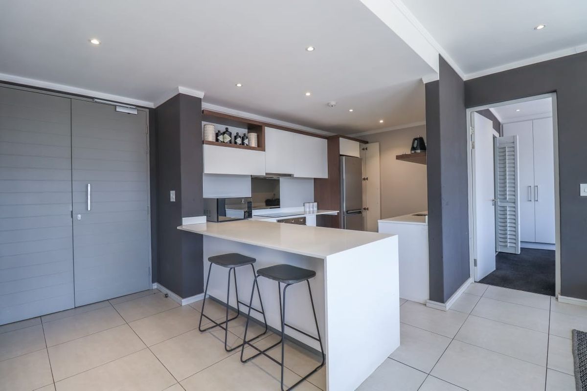 Modern kitchen featuring sleek white cabinetry, stone countertops, a breakfast bar, and recessed lighting, seamlessly integrated into an open-plan living space.