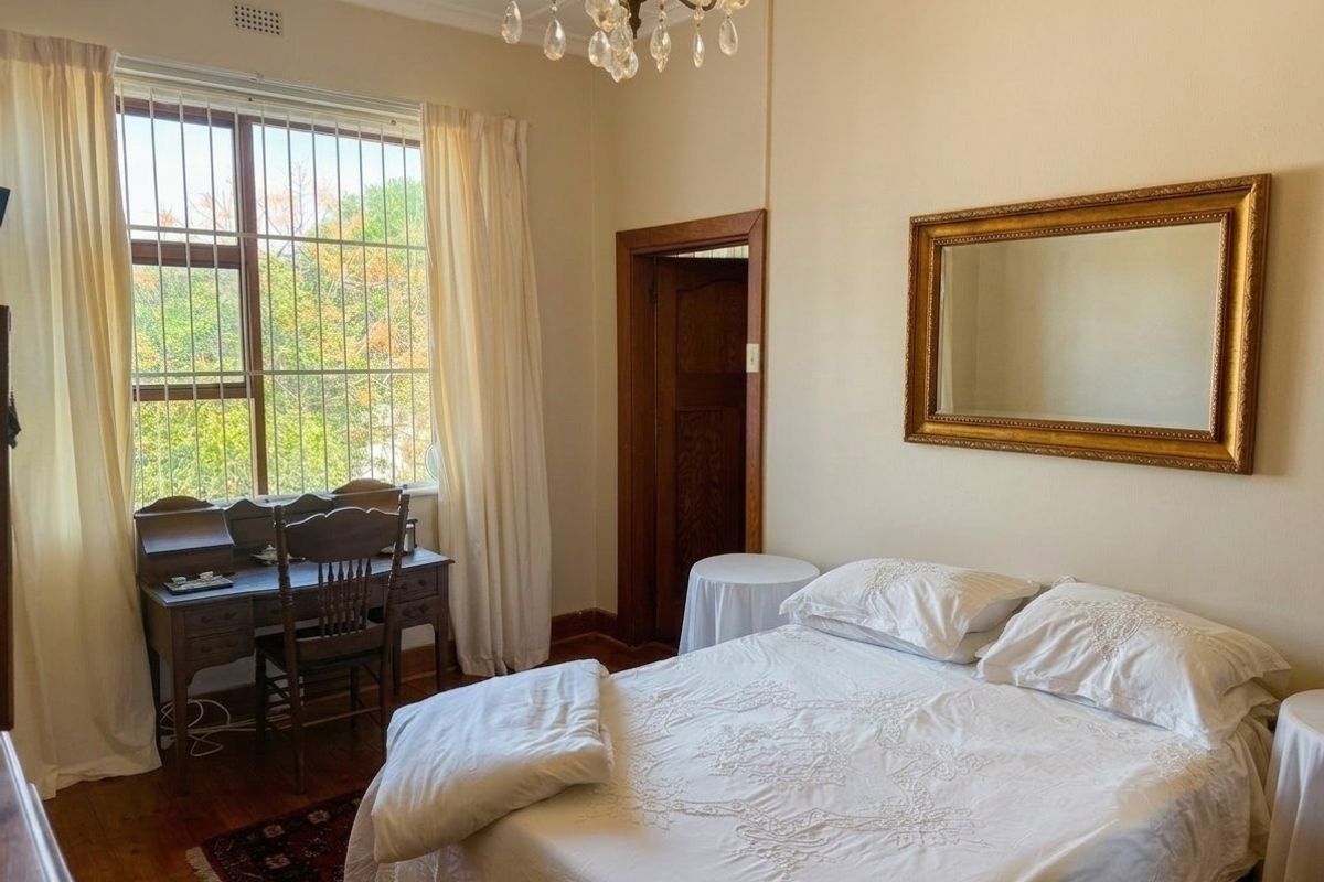 A well-lit bedroom featuring original hardwood flooring, a classic crystal chandelier, and large windows fitted with security bars.