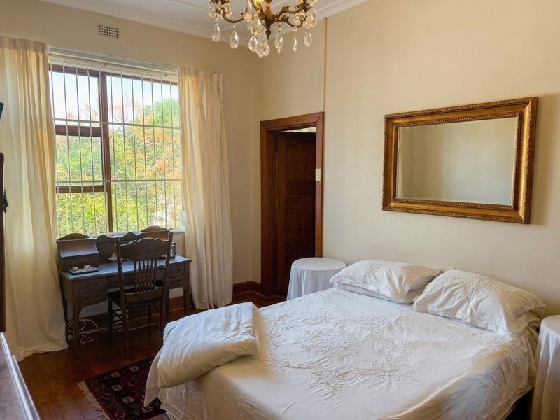 A well-lit bedroom featuring original hardwood flooring, a classic crystal chandelier, and large windows fitted with security bars.