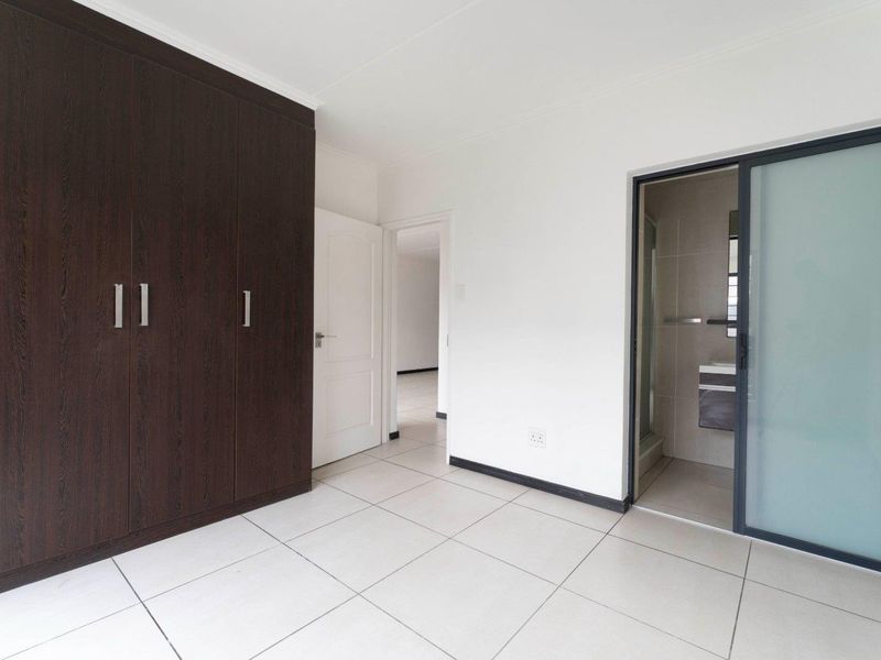 A clean, modern bedroom featuring large dark wood built-in cupboards, neutral tiled flooring, and direct access to an en-suite bathroom.