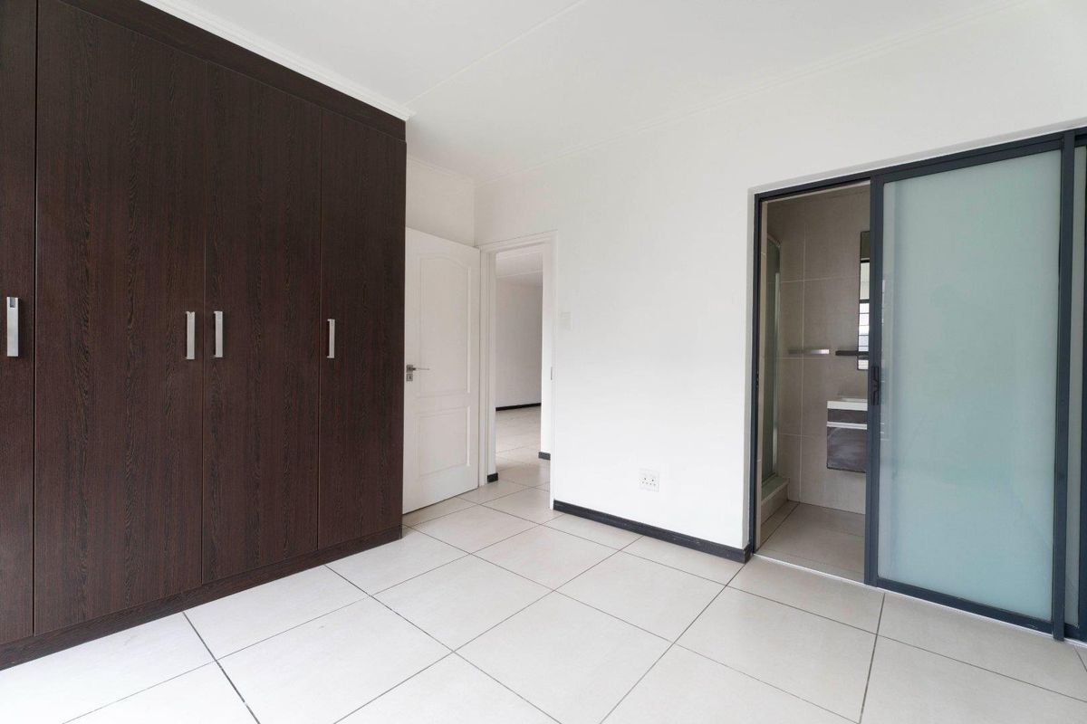 A clean, modern bedroom featuring large dark wood built-in cupboards, neutral tiled flooring, and direct access to an en-suite bathroom.