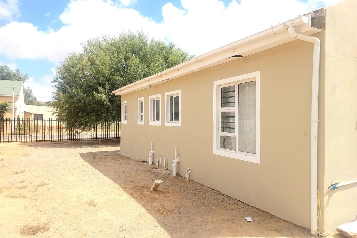 Rear exterior view of the property showing the building facade, window installations, guttering system, and exposed plumbing pipes on a sandy ground surface.