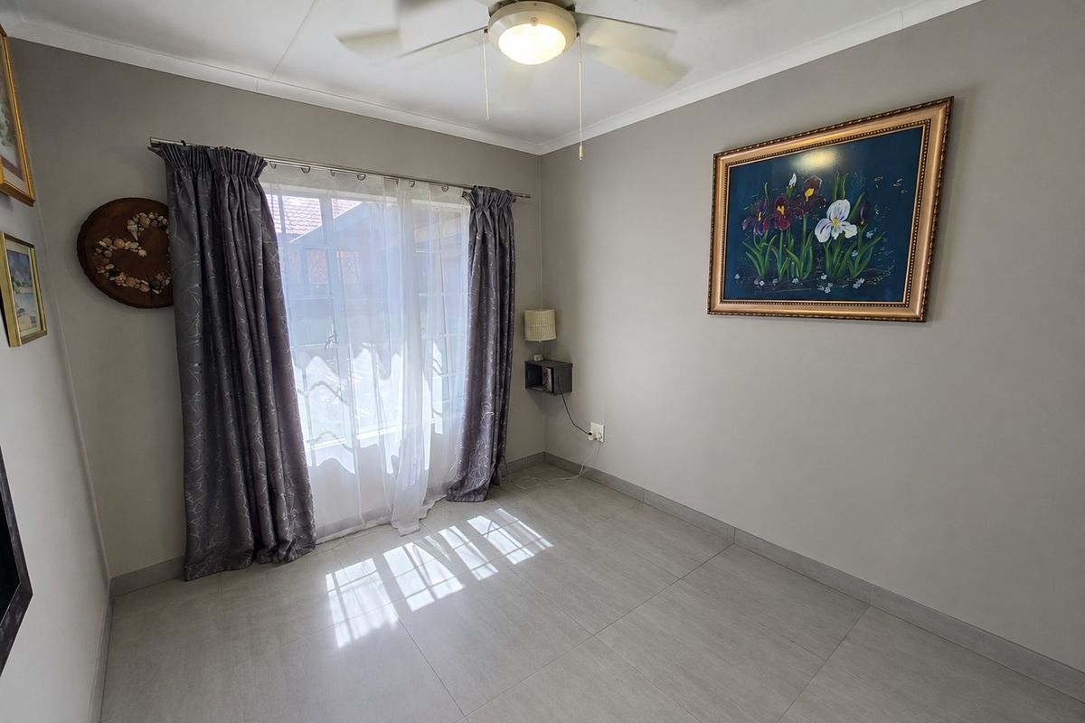 A well-maintained bedroom featuring neutral tiled flooring, a ceiling fan, and large windows with curtains, offering a clean and bright living space.