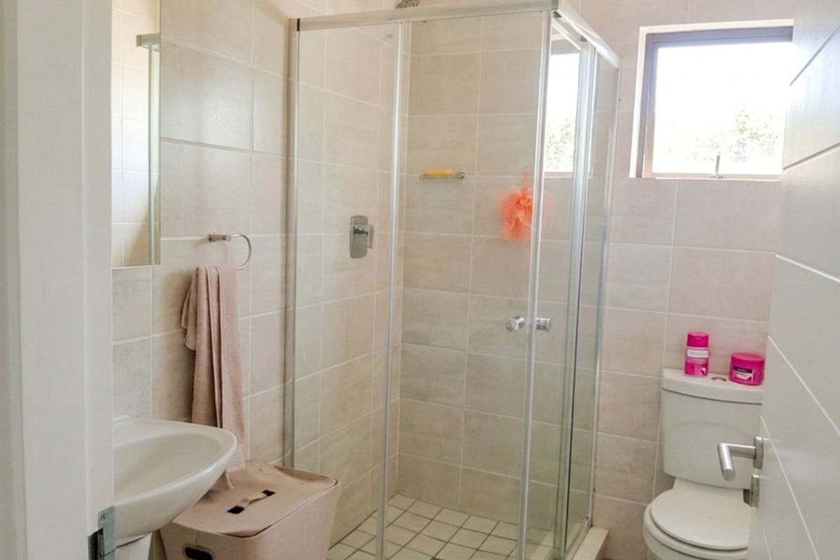 A functional bathroom featuring a glass-enclosed shower, pedestal basin, and toilet with neutral wall and floor tiling.