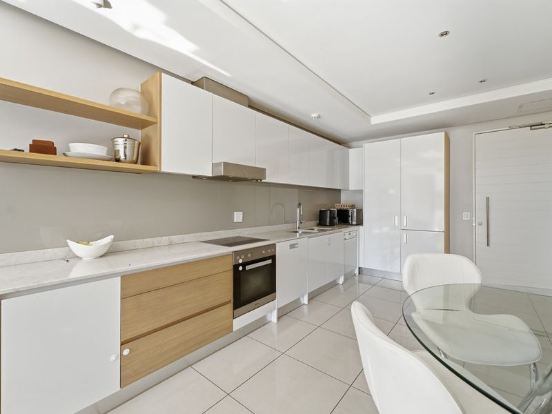 Modern open-plan kitchen featuring sleek white and wood-tone cabinetry, stone countertops, integrated appliances, and polished floor tiles in a bright, contemporary apartment setting.