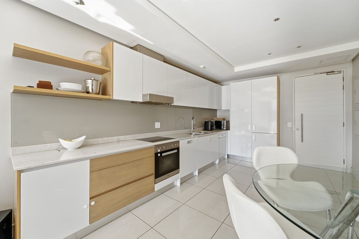 Modern open-plan kitchen featuring sleek white and wood-tone cabinetry, stone countertops, integrated appliances, and polished floor tiles in a bright, contemporary apartment setting.