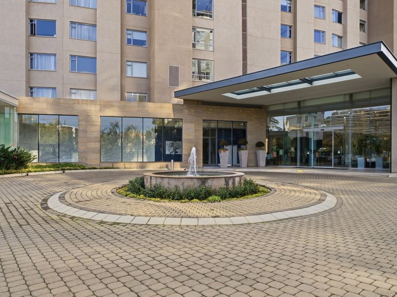 A modern apartment building entrance featuring a paved circular driveway, a central stone fountain, and a sleek glass canopy over the main entrance.