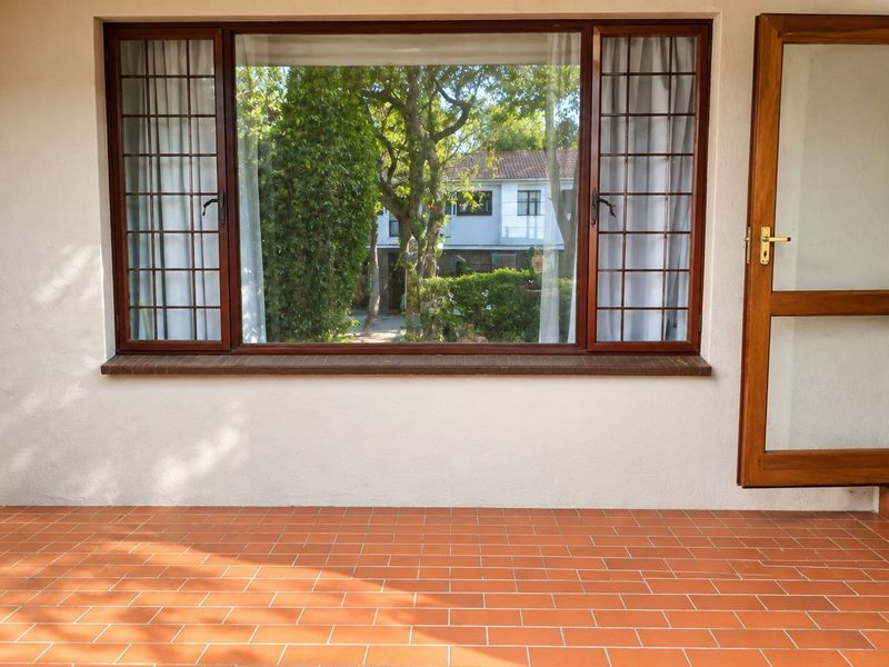 A covered patio area featuring terracotta tiled flooring, a large timber-framed window with security bars, and a timber door leading to the interior.