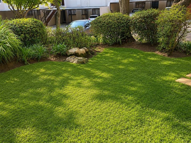A well-maintained private garden area featuring a lush green lawn, established shrubbery, and stone pathway accents within an apartment complex setting.