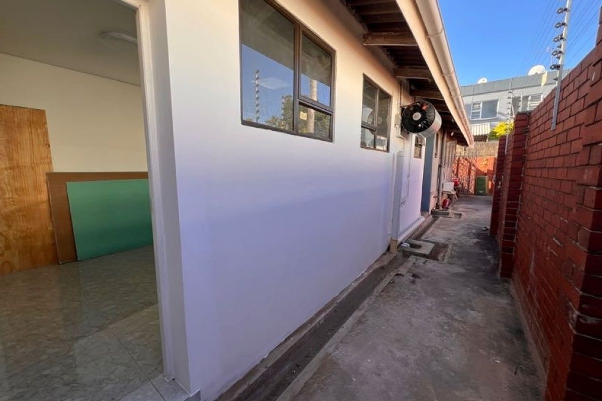 A side exterior view of the property featuring a concrete pathway, brick boundary wall, and aluminum window frames.